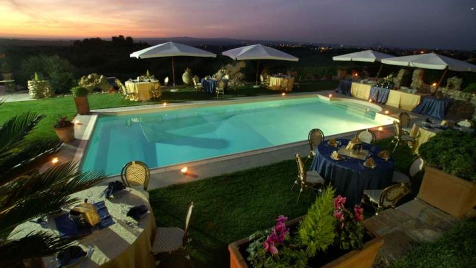 Piscina rettangolare all’aperto illuminata al crepuscolo, circondata da prato e da tavoli apparecchiati con tovaglie e sedie. Attorno ci sono ombrelloni bianchi, fioriere e muretti in pietra, con vista su una campagna collinare sullo sfondo.