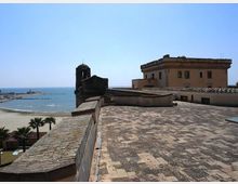 Terrazza in pietra su una fortificazione costiera, con muretto di cinta e edificio storico con torre campanaria; sullo sfondo si vedono la spiaggia e il mare. A destra compare un edificio rettangolare intonacato con tetto basso e finestre affacciate verso la costa.