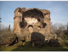 Rovine di un edificio antico in mattoni con grande abside semicircolare e nicchie ad arco, parzialmente crollato, circondato da massi e resti di murature. Sullo sfondo si vedono alberi spogli e cielo sereno.