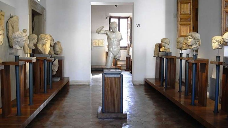Sala museale con pareti bianche e pavimento in pietra, con una statua classica al centro e busti in marmo esposti su supporti in legno e metallo lungo entrambi i lati. Sul fondo si vede un’apertura verso un’altra stanza con una finestra e una porta in legno.