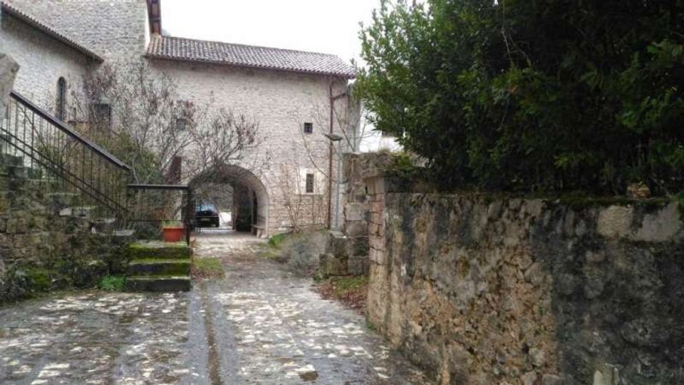 Cortile in pietra con pavimentazione acciottolata che conduce a un arco d’ingresso in un edificio in muratura, con tetto a coppi. Ai lati si vedono un muretto in pietra con vegetazione fitta e una scala esterna con ringhiera metallica.