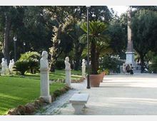 Viale pedonale in un parco con ampi prati e alberi alti, fiancheggiato da statue e busti in pietra su piedistalli. Lungo il percorso si vedono lampioni, una panchina, grandi vasi con palme e un alto monumento/obelisco sullo sfondo.