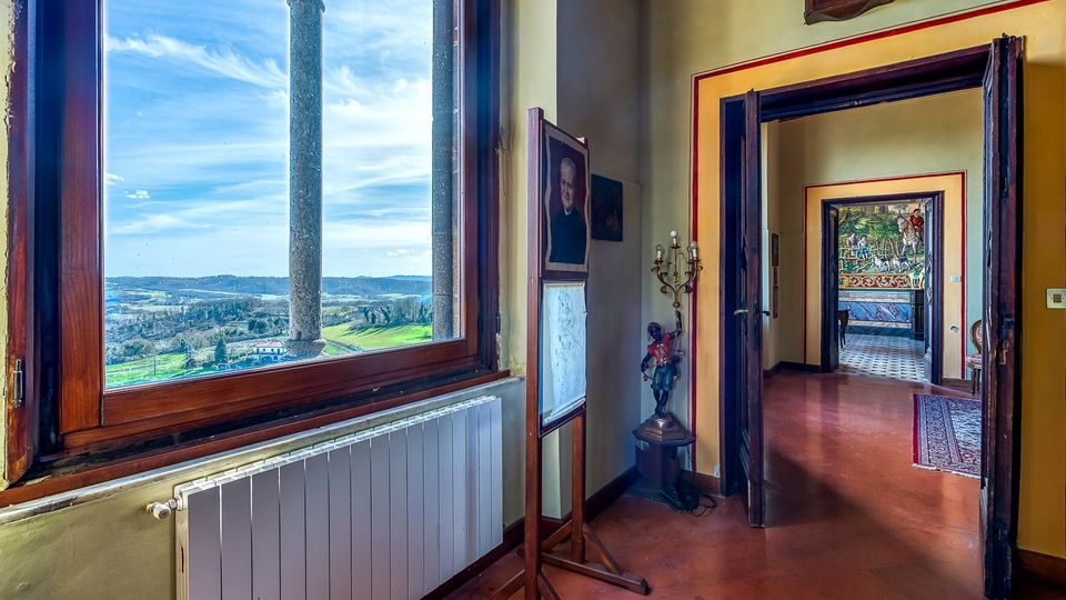 Interno di un corridoio in edificio storico con pavimento in cotto, porte in legno e pareti dai toni chiari, illuminato da una grande finestra ad arco con colonna in pietra che affaccia su colline e campagna. In fondo si vede una stanza con affresco murale e pavimento a piastrelle, con lampada decorativa e piccoli elementi d’arredo lungo il passaggio.