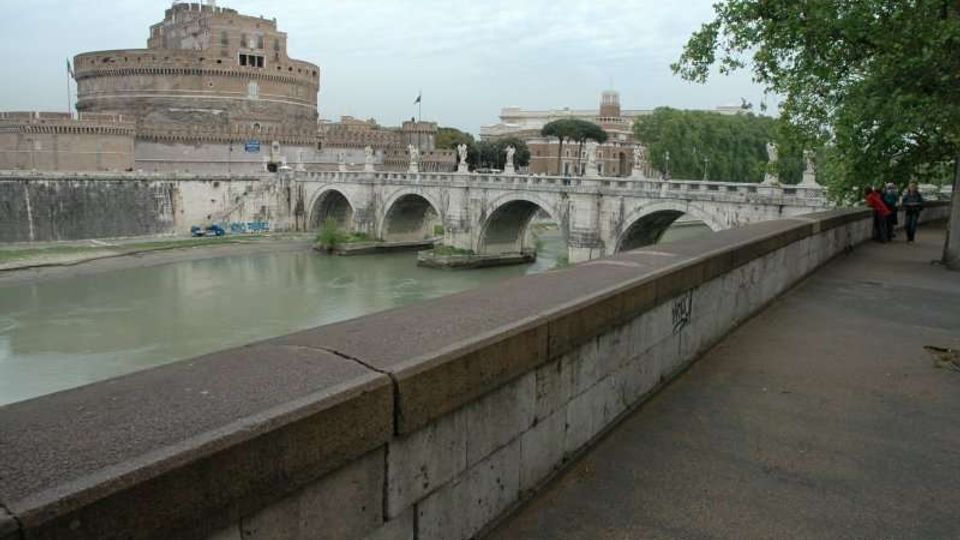 Veduta del Tevere con un ponte in pietra a più arcate e, sullo sfondo, una grande fortezza cilindrica in muratura. In primo piano si vede una lunga banchina con muro in pietra e un viale alberato lungo il fiume.
