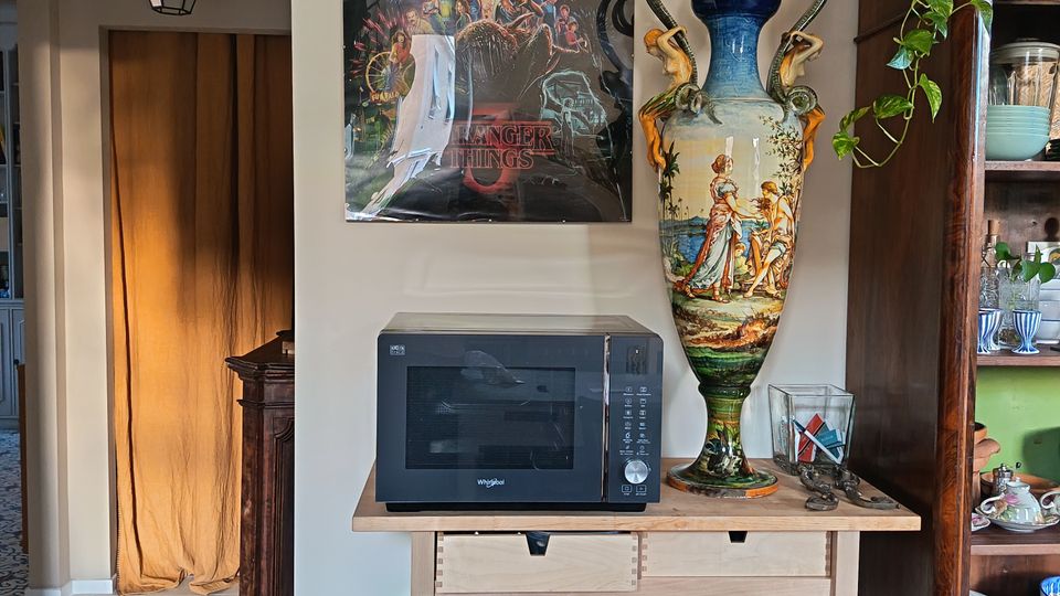 Angolo di cucina con carrellino in legno che sostiene un forno a microonde e una grande anfora decorativa, sotto un poster appeso alla parete. A destra c’è una credenza in legno con stoviglie e una pianta ricadente, mentre a sinistra si vede un passaggio con tenda color ocra e lanterna a soffitto.