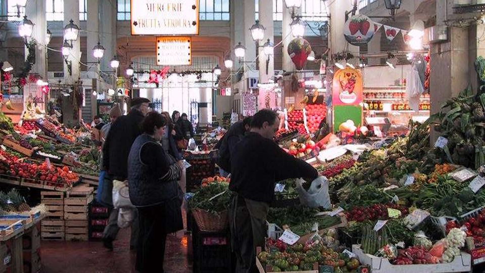 Interno di un mercato coperto con alte colonne bianche, soffitto a travi e file di lampioni sospesi lungo il corridoio centrale. Banchi su entrambi i lati espongono frutta e verdura in cassette, con cartelli e un’insegna “PESCHERIE” visibile sullo sfondo.