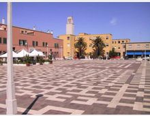 Ampia piazza urbana pavimentata con motivi geometrici in pietra chiara e scura, circondata da edifici bassi color ocra e rosso. Sullo sfondo si vedono una torre campanaria, alcune palme e strutture con tende bianche lungo il lato sinistro.