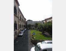 Cortile interno tra edifici in stile storico con facciate chiare e finestre ad arco con persiane, affacciato su una strada stretta con auto parcheggiate. Al centro c’è un’area verde con alberi e un cartello di parcheggio, chiusa sul fondo da un edificio moderno con finestre verticali.