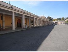 Cortile asfaltato di un complesso a un piano con un lungo porticato laterale sostenuto da colonne rettangolari e pareti color ocra. Sul cornicione corre una scritta e, sullo sfondo, si vedono altri edifici bassi con alcune alberature.