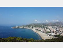 Veduta panoramica dall’alto di una grande baia costiera con lunga spiaggia sabbiosa e mare calmo, affiancata da un centro abitato denso lungo il litorale. Sullo sfondo si vedono colline e montagne, con vegetazione in primo piano e cielo sereno.