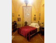 Camera da letto in stile tradizionale con soffitto a volta dipinto, pareti gialle decorate e pavimento in cotto. Un letto con coperta bordeaux è al centro sotto una finestra alta con tenda in pizzo, affiancato da comodini e quadri appesi alle pareti.