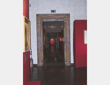 Interno di museo con pareti rosse e pavimento in legno scuro lucido, visto attraverso un grande portale rettangolare con cornice in marmo. In fondo si apre un lungo corridoio in prospettiva con più passaggi allineati e quadri esposti alle pareti, illuminato da luci a soffitto.