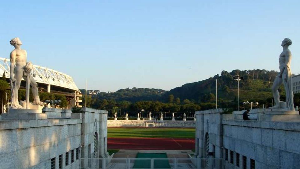 Ingresso monumentale di uno stadio con due statue in pietra ai lati e gradinate in marmo che conducono a una pista di atletica rossa e al campo centrale. Sullo sfondo si vedono colline verdi e una tribuna con struttura metallica sotto un cielo sereno.