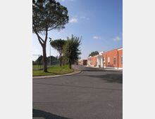 Strada asfaltata curva in un’area urbana tranquilla, con alberi alti e un’area verde recintata a sinistra. A destra si vedono edifici bassi color rosso mattone con ingressi bianchi e finestre allineate lungo il marciapiede.