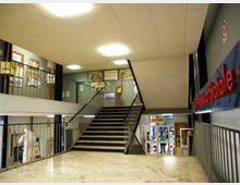 Atrio interno di un edificio pubblico con pavimento in marmo e un’ampia scalinata centrale con corrimano in metallo che conduce al piano superiore. Lungo le pareti e sui ballatoi sono visibili pannelli espositivi e una scritta a parete, con luci a soffitto e accesso a una sala sul fondo.