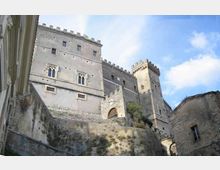 Veduta dal basso di una fortezza in pietra su una collina, con alte mura e torri merlate e piccole finestre. In primo piano si vedono i muri di contenimento e un edificio in pietra, con cielo azzurro sullo sfondo.
