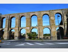 Antico acquedotto in pietra con due ordini di grandi arcate, che attraversa una strada asfaltata con segnaletica orizzontale. Sullo sfondo si vedono lampioni, una ringhiera e alberi sotto un cielo sereno.