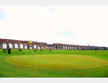 Campo da golf con green e bandierina in primo piano, circondato da ampie distese erbose. Sullo sfondo si estende un lungo acquedotto in mattoni con una serie di arcate, sotto un cielo chiaro.