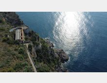 Veduta aerea di una costa rocciosa con scogliere e vegetazione mediterranea, affiancata da una strada stretta che conduce a un edificio rettangolare vicino al bordo del promontorio. Il mare occupa la maggior parte dell’immagine, con riflessi di luce sulla superficie dell’acqua.