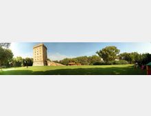 Ampio prato verde in un parco con una torre in pietra alta e rettangolare, affiancata da un basso edificio con tetto rosso. Sullo sfondo si vede una fila di alberi sotto un cielo sereno.