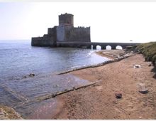 Castello costiero in pietra su un isolotto, collegato alla riva da un ponte basso con arcate, affacciato sul mare calmo. In primo piano si vede una spiaggia sabbiosa con scogliera e vegetazione bassa lungo il margine.