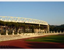 Vista di uno stadio con struttura superiore in traliccio bianco, affiancato da una pista di atletica rossa e un campo erboso. In primo piano si vedono gradinate basse con statue lungo il perimetro e una fila di pini, con colline sullo sfondo.