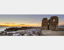 Rovine in pietra di un’antica struttura costiera con due muri parzialmente crollati e aperture, su una spiaggia rocciosa. In primo piano una scogliera di massi conduce al mare calmo, con il sole al tramonto all’orizzonte.