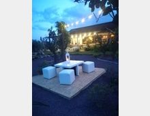 Area lounge all’aperto in giardino al crepuscolo, con tavolino bianco e quattro pouf su una pedana e una lanterna al centro. Sullo sfondo si vede un edificio con veranda illuminata e fili di lucine sospese tra gli alberi.