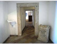 Corridoio interno con pareti bianche e pavimento in pietra, attraversato da un grande portale rettangolare in pietra che conduce a un’altra sala. Ai lati si vedono un leggio trasparente e un basamento in pietra con iscrizione, in un ambiente dall’aspetto museale.