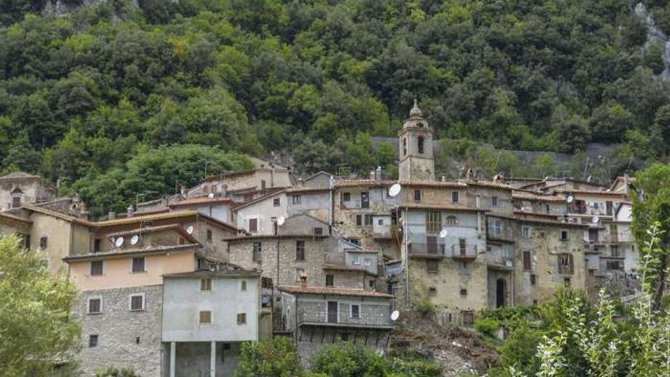 Borgo collinare con case in pietra e intonaco addossate al pendio, tetti in coppi e una piccola torre campanaria al centro. Sullo sfondo si vede un fitto bosco montano con affioramenti rocciosi.
