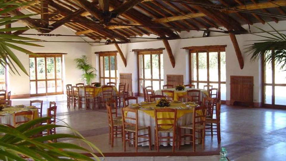 Ampia sala per ricevimenti con soffitto alto in travi di legno a vista e grandi finestre e porte vetrate che lasciano entrare molta luce. Tavoli rotondi apparecchiati con tovaglie bianche e gialle e sedie in legno su pavimento in pietra, con piante decorative ai lati.