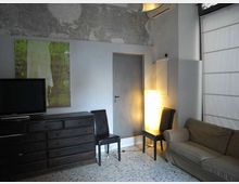 Interno di una stanza in stile moderno-industriale con pareti chiare e una fascia di intonaco grezzo vicino al soffitto. Sono visibili una cassettiera scura con TV e quadro, una porta centrale, due sedie nere, una lampada da terra luminosa e un divano accanto a una grande finestra con tenda.