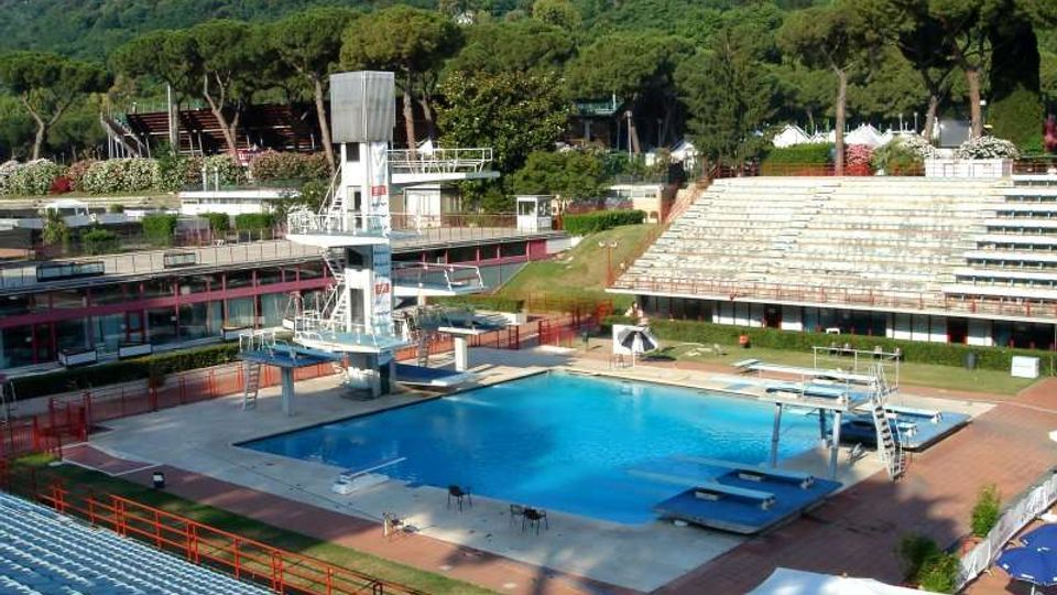 Piscina all’aperto in un complesso sportivo con vasca rettangolare azzurra, piattaforme e torre per tuffi, circondata da bordo pavimentato e ringhiere. Intorno si vedono gradinate tipo stadio e una collina boscosa sullo sfondo.