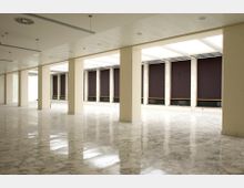 Ampio spazio interno vuoto, simile a una sala o open space, con colonne rettangolari in fila, pavimento in marmo lucido e soffitto con pannelli e illuminazione incassata. Lungo un lato si vedono grandi finestre schermate da pannelli scuri e lucernari che lasciano entrare luce naturale.