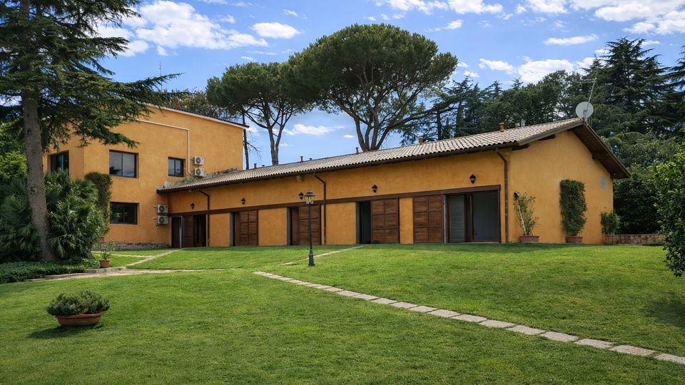 Edificio basso in intonaco giallo con tetto a falde e grandi porte in legno, affacciato su un ampio prato con vialetto in pietra e lampione. Sullo sfondo si vedono alti alberi mediterranei e un cielo azzurro con nuvole sparse.