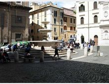 Piazza lastricata in sampietrini con una grande fontana in pietra al centro e gradini tutt’intorno. Sullo sfondo si vedono edifici storici a più piani con persiane e facciate chiare, oltre a una facciata di chiesa e alcuni dissuasori in pietra lungo il bordo della piazza.