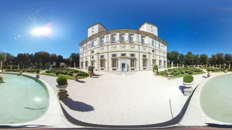 Veduta panoramica di un palazzo storico a pianta curva, con facciata chiara e finestre regolari, affacciato su un ampio viale in ghiaia. In primo piano ci sono due vasche d’acqua e, tutt’intorno, un giardino formale con siepi geometriche, statue e alberi sullo sfondo.