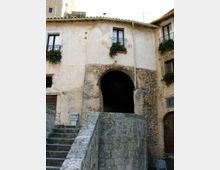 Facciata di un edificio storico in pietra e intonaco con un grande arco d’ingresso che conduce a un passaggio coperto. In primo piano si vedono una scalinata e un muretto in pietra, con finestre e piccoli balconi con ringhiere in ferro e fioriere.
