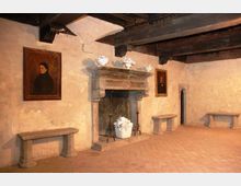 Interno di una sala storica con grande camino in pietra al centro, soffitto con travi di legno a vista e pavimento in cotto. Alle pareti intonacate sono appesi ritratti incorniciati e sono presenti panche in pietra lungo i lati.