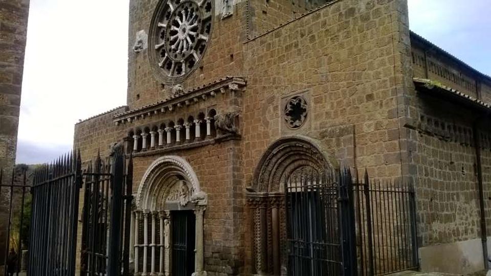 Facciata in pietra di una chiesa medievale con grande rosone circolare, portali ad arco decorati e piccoli colonnati scolpiti. Davanti all’ingresso sono presenti alte cancellate e recinzioni in ferro battuto.