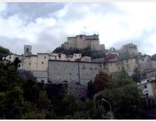 Borgo collinare con case in pietra addossate al pendio e un grande castello o fortezza sulla sommità. In primo piano si vede vegetazione fitta, mentre sullo sfondo il cielo è nuvoloso.