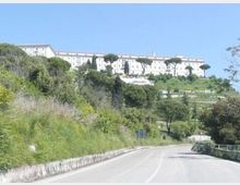 Strada asfaltata in curva che attraversa una collina verde con vegetazione mediterranea e alberi ad alto fusto. In cima al pendio si vede un grande edificio chiaro e rettangolare con più piani, circondato da terrazze e alberi, sotto un cielo sereno.