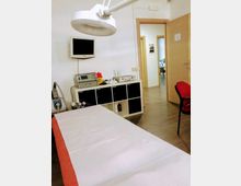 Ambulatorio medico con lettino da visita coperto da lenzuolo, lampada chirurgica a soffitto e monitor a parete sopra un mobile con attrezzature. Porta aperta verso un corridoio interno e pavimento in linoleum chiaro.