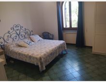 Camera da letto con letto matrimoniale e testiera in ferro battuto blu, copriletto chiaro a motivi marini e pavimento in piastrelle verdi. Una grande finestra ad arco con tende scure illumina la stanza; vicino ci sono una sedia e un armadio bianco.