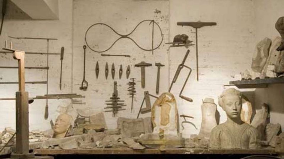 Interno di un laboratorio di scultura con parete bianca su cui sono appesi utensili e sagome metalliche. In primo piano un banco da lavoro ingombro di frammenti e busti in gesso, con scaffali laterali pieni di modelli e materiali.