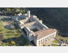 Veduta aerea di un grande complesso monastico in pietra con tetti in coppi, organizzato attorno a chiostri e cortili interni, con un campanile quadrato centrale. L’edificio è circondato da aree verdi e si trova ai margini di un bosco su un pendio.