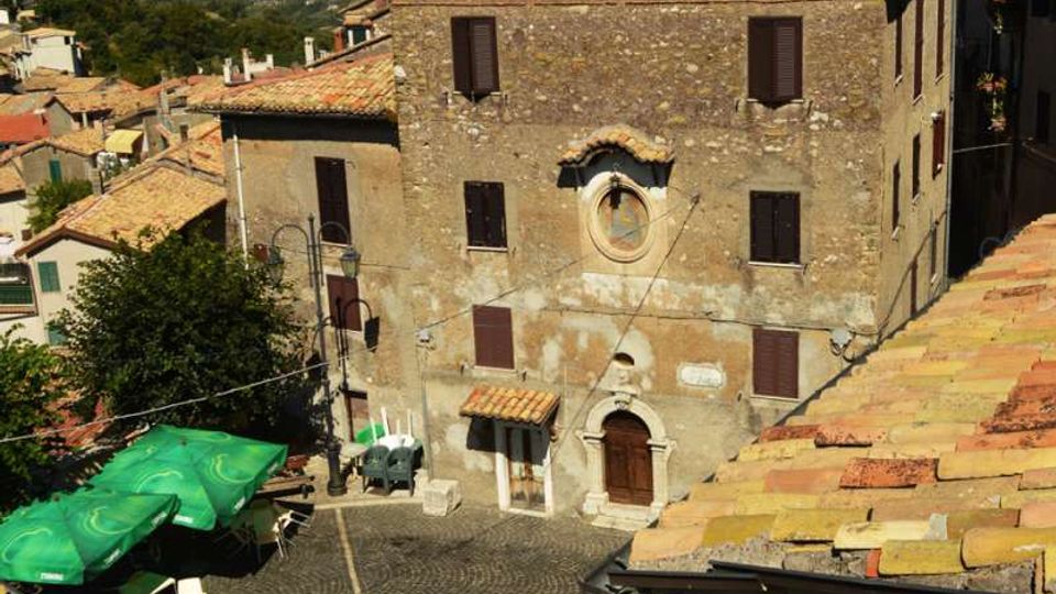 Piazza lastricata in pietra con disegno circolare, circondata da edifici in muratura con tetti in coppi; in primo piano un grande palazzo in pietra con persiane scure e portale ad arco. Sullo sfondo si vedono colline e montagne sotto un cielo sereno.