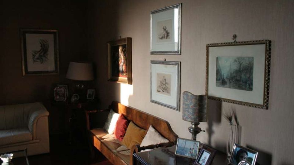 Salotto interno con divano in legno e cuscini, poltrona chiara e pareti decorate con quadri incorniciati. In primo piano un mobile con cornici e una lampada da tavolo, illuminati da luce naturale proveniente da una finestra fuori campo.
