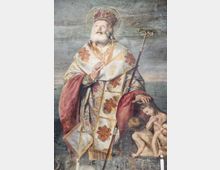 Affresco religioso su parete in pietra raffigurante un santo con aureola e vesti liturgiche riccamente decorate, con pastorale e mano alzata in gesto di benedizione. Ai suoi piedi è visibile una piccola scena con figure infantili, dipinta nello stesso riquadro murale.