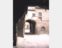 Passaggio ad arco in pietra con scalinate laterali e facciate in muratura con finestre; sotto l’arco si intravede un portone rosso in fondo a una piccola piazza lastricata. Ai lati sono presenti un lampione e un segnale stradale di divieto di sosta.